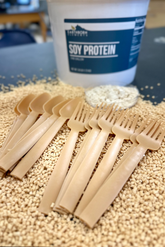 Beige disposable forks and spoons made from soy protein arranged on a bed of soy pellets, with a container labeled “Soy Protein” in the background